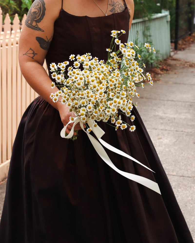 Person in a dark brown dress holding a bouquet of white daisies with a white ribbon.