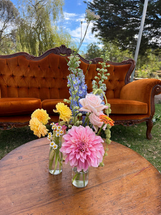 Floral arrangement in small bottles on a wooden table with an orange sofa and greenery in the background