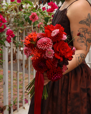 Person holding a bouquet of red and pink flowers with tattoos visible on arms, against a garden background.