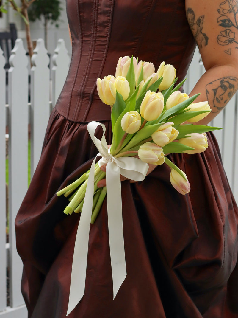 Bride holding a bouquet of yellow tulips with a white ribbon in front of a white picket fence.