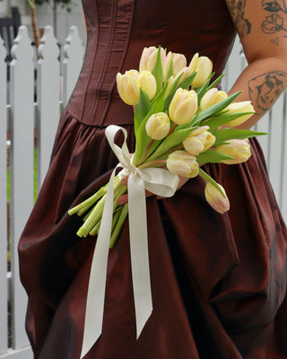 Person wearing a brown dress holding a bouquet of yellow tulips with a white ribbon.